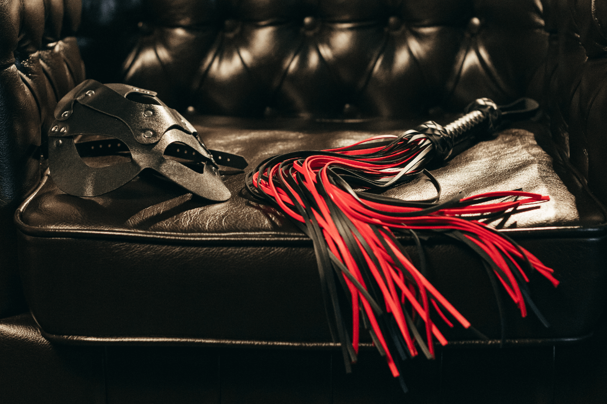 BDSM Mythen Symbolbild: rote Peitsche und Ledermaske auf Ledercouch BDSM myths Symbolic image: red whip and leather mask on leather couch