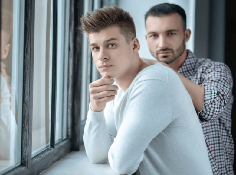Vertrauensbruch in der Beziehung. Symbolbild: Zwei Männer stehen ernst dreinblickend an einem Fenster Breach of trust in a relationship. Symbolic image: Two men standing at a window looking serious