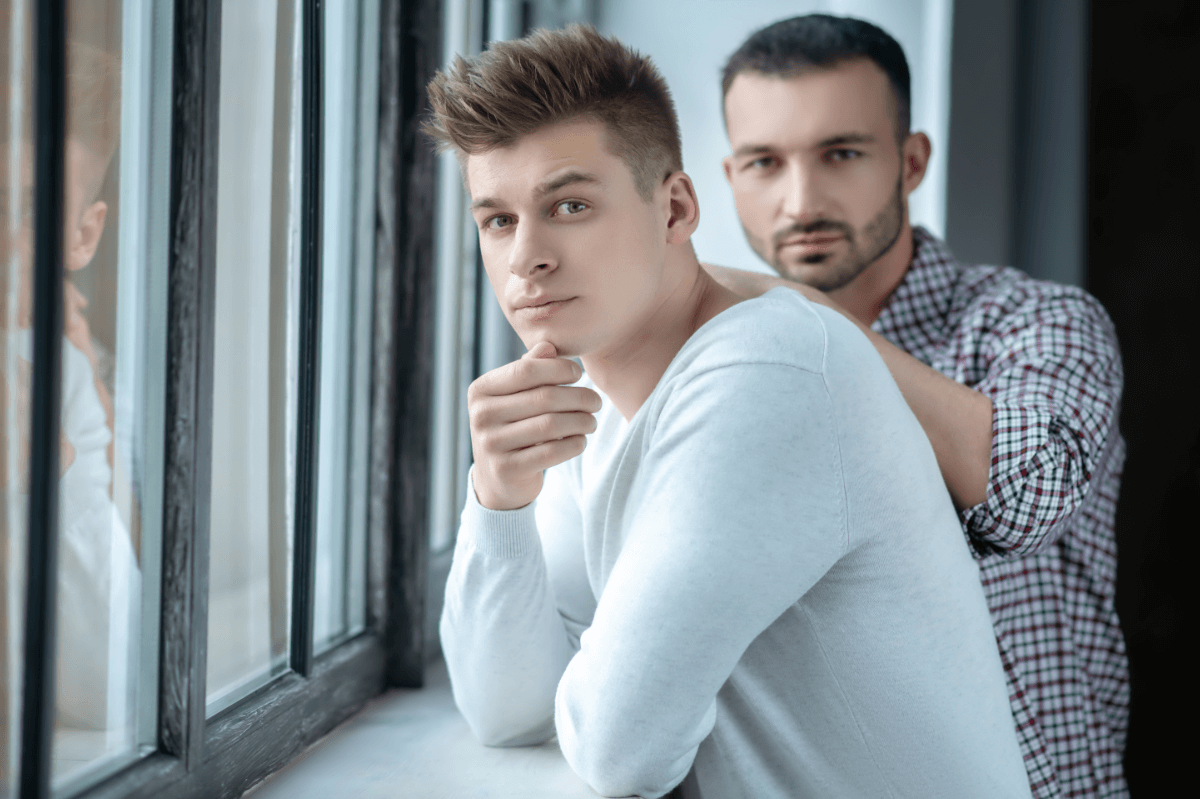 Vertrauensbruch in der Beziehung. Symbolbild: Zwei Männer stehen ernst dreinblickend an einem Fenster Breach of trust in a relationship. Symbolic image: Two men standing at a window looking serious