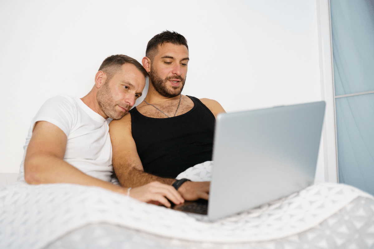 Beziehungsprobleme schwule Paare. Symbolbild: Zwei Männer liegen im Bett und schauen gemeinsam in den Laptop. Englisch: Relationship problems of gay couples. Symbolic image: Two men lying in bed and looking at a laptop together.