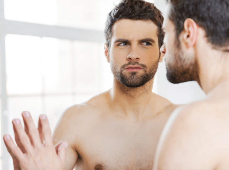 Confidence beim Sex - Symbolbild: Ein Mann mit nacktem Oberkörper schaut kritisch in den Spiegel. Sexual Confidence – symbolic image: a man with a naked torso looks critically in the mirror.