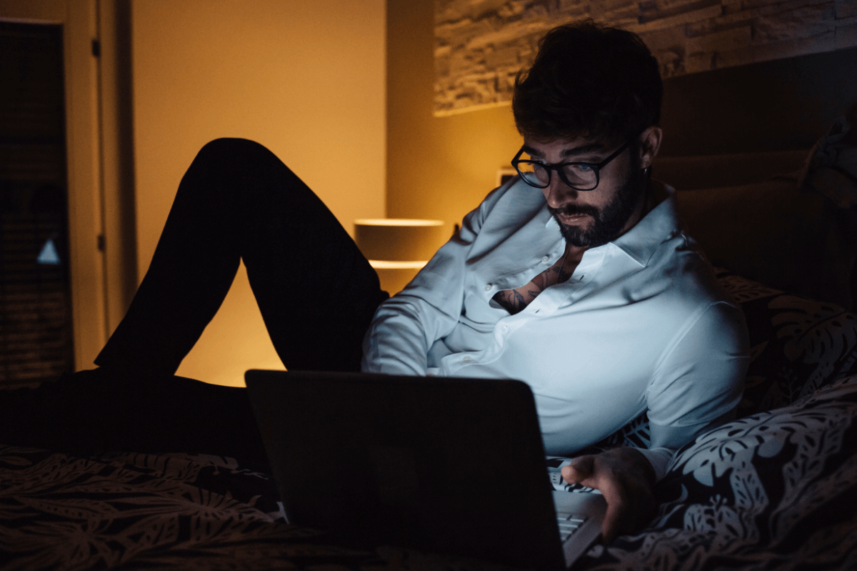 Pornosucht. Symbolbild: Ein Mann liegt im Halbdunkeln vor seinem Laptop auf dem Bett. Englisch: Porn addiction. Symbolic image: a man is lying on his bed in front of his laptop in semi-darkness.