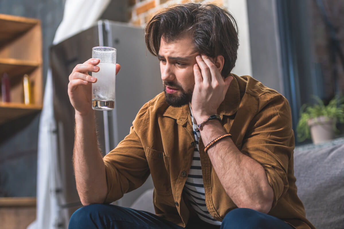 Poppers Nebenwirkungen Symbolbild: Ein Mann hält sich den Kopf vor Schmerzen und hält ein Glas mit Schmerzmittel in der Hand. Englisch: Side effects of poppers Symbolic image: A man is holding his head in pain and holding a glass of painkillers in his hand.