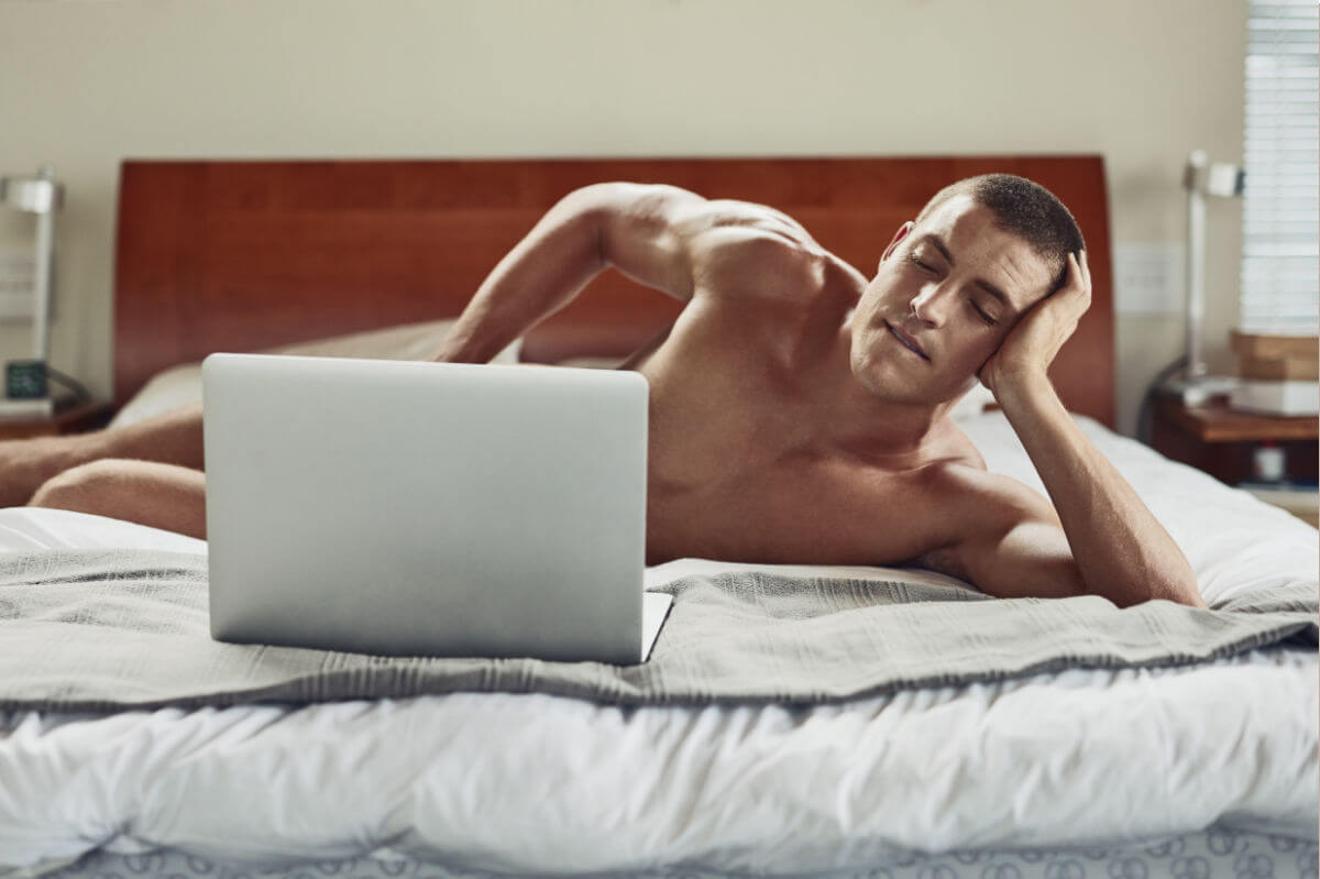 Pornosexuell Symbolbild: Ein Mann liegt nackt auf dem Bett und schaut in einen Laptop, der sein Geschlechtsteil verdeckt. Englisch: Pornosexual symbol: a man lies naked on the bed and looks at a laptop that covers his genitals.