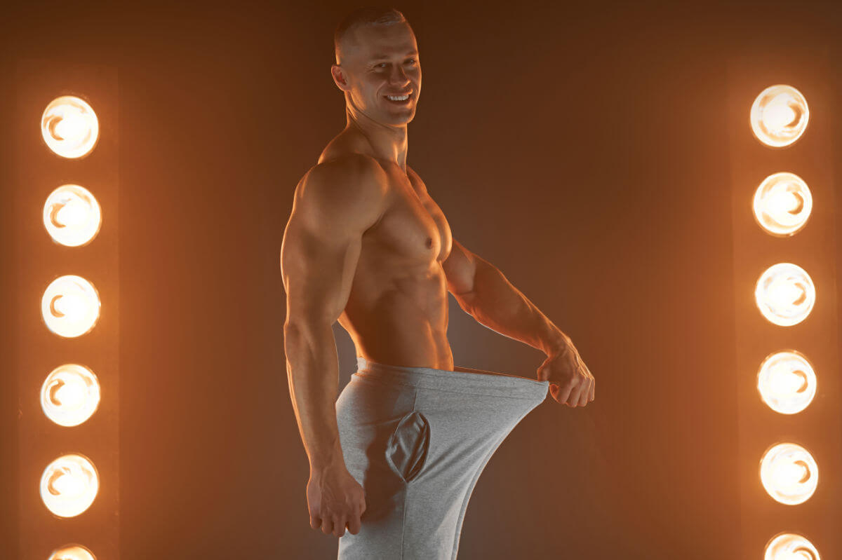 Penislänge Symbolbild: Ein Mann mit nacktem Oberkörper hält seine Hose im Schritt soweit von sich weg, dass ein langer Penis angedeutet wird. Englisch: Penis length Symbolic image: A man with a bare upper body holds his pants away from his crotch, suggesting a long penis.