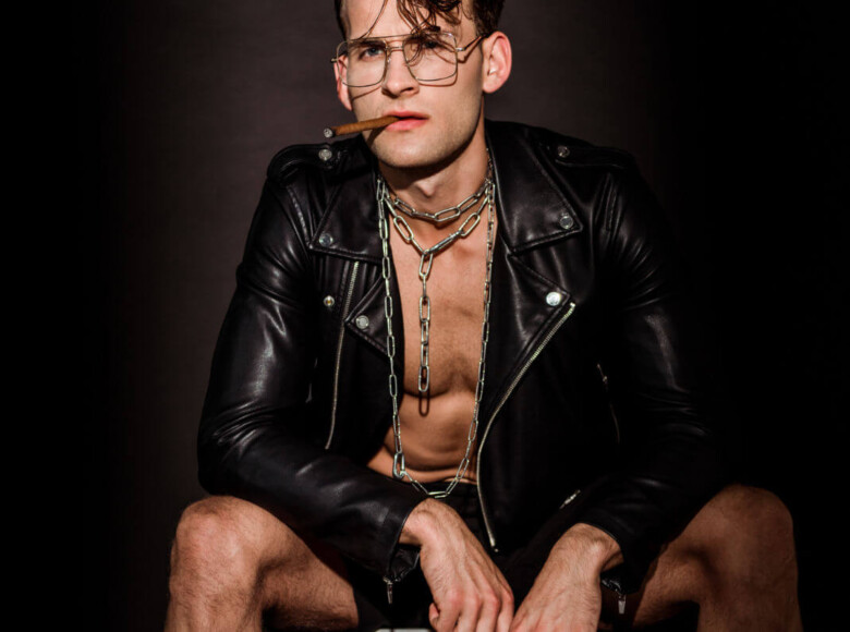 Fetisch vs. Kink Symbolbild: Ein Mann mit offener Lederjacke und Zigarre im Mund schaut lassziv in die Kamera. Fetish vs. Kink Symbolic image: A man with an open leather jacket and a cigar in his mouth looks lasciviously into the camera.