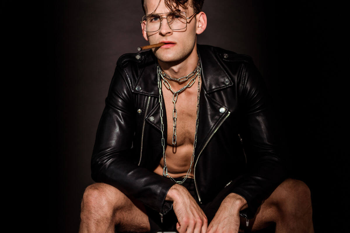 Fetisch vs. Kink Symbolbild: Ein Mann mit offener Lederjacke und Zigarre im Mund schaut lassziv in die Kamera. Fetish vs. Kink Symbolic image: A man with an open leather jacket and a cigar in his mouth looks lasciviously into the camera.