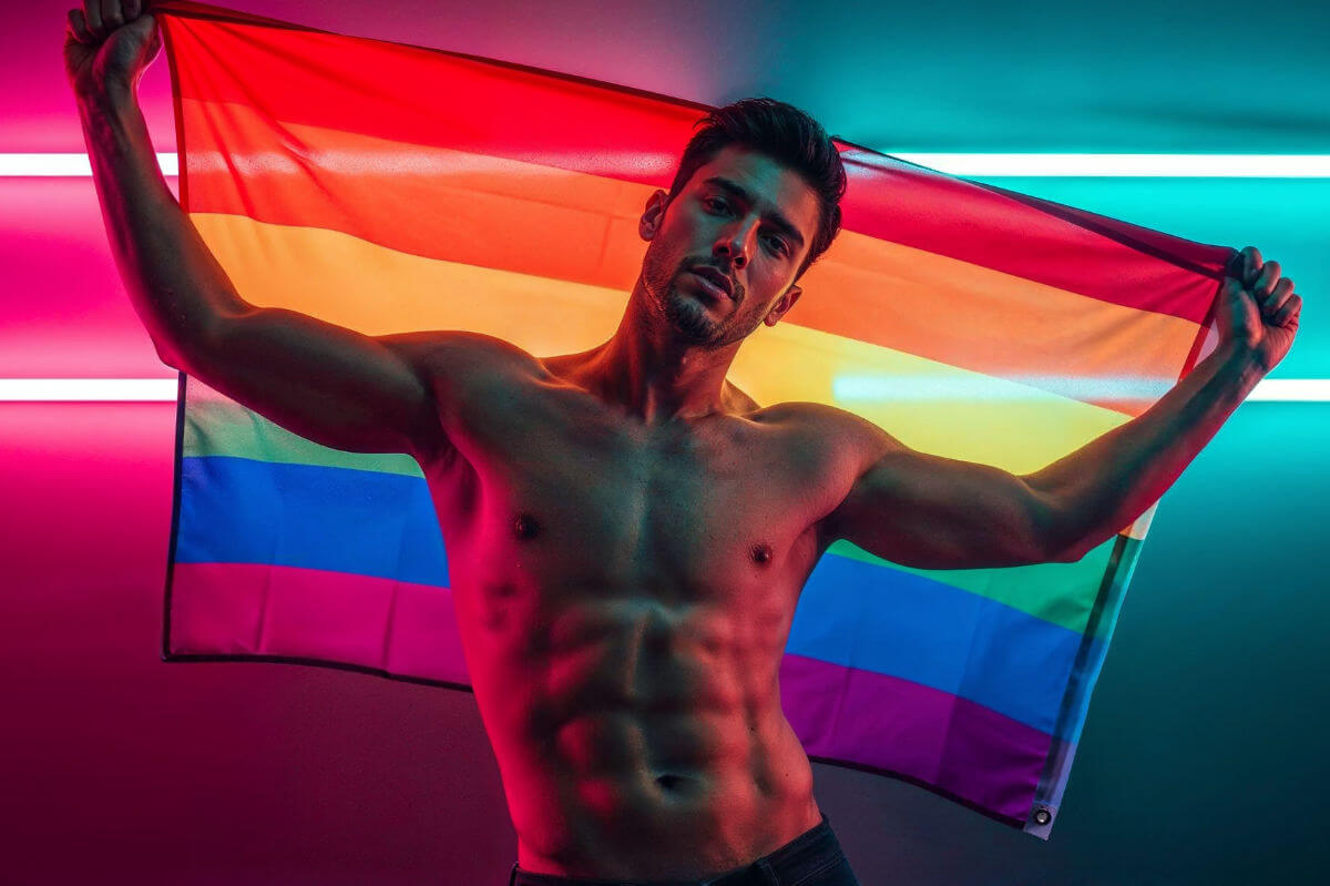 CSD 2025 Termine Juli, August & September. Symbolbild: Ein Mann mit nacktem Oberkörper hält die Regenbogenfahne. CSD 2025 dates for July, August, and September. Symbolic image: A man with a bare torso holding a rainbow flag.