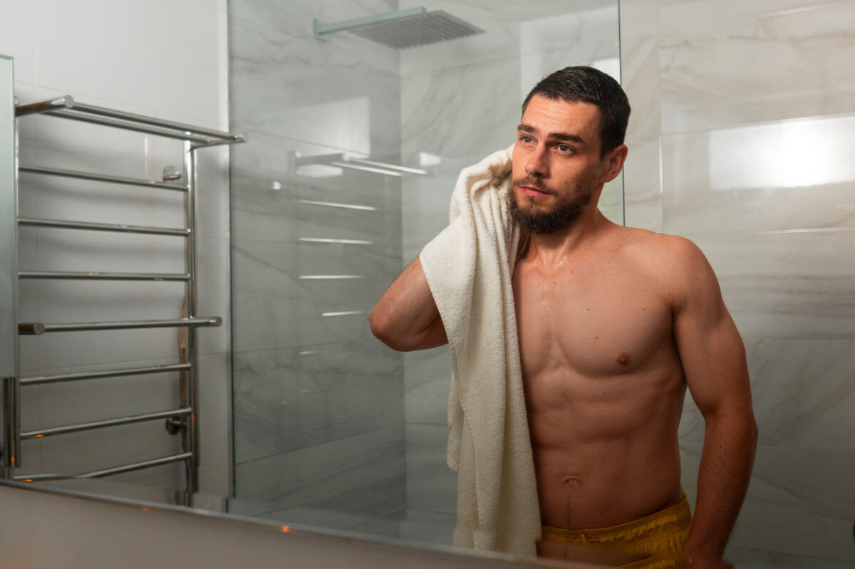 Aftercare für Tops und Doms Symbolbild Topdrop: Ein Mann steht nackt vor dem Badezimmerspiegel und trocknet sich mit einem Handtuch ab. . Aftercare for tops and doms Symbolic image of a top drop: A man stands naked in front of the bathroom mirror and dries himself with a towel.