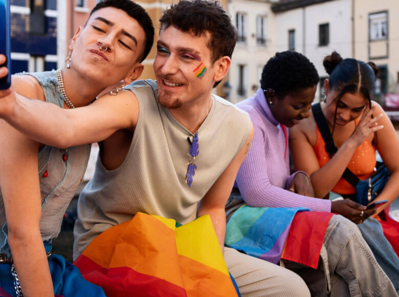 Erfahrungen queerer Menschen in Schule und Ausbildung Symbolbild: Zwei männliche Teenager sowie zwei weibliche sitzen auf einer Bank und halten die Regenbogen-Flagge auf den Beinen. Die beiden Jungs machen ein Selfie. Die Mädchen schauen gemeinsam in ein Handy. . Experiences of queer people in school and education Symbolic image: Two male teenagers and two female teenagers are sitting on a bench holding a rainbow flag between them. The two boys are taking a selfie. The girls are looking at a cell phone together.