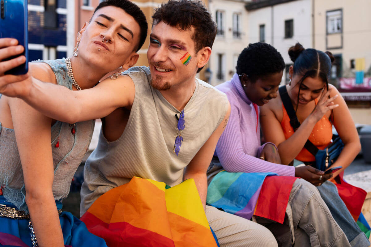 Erfahrungen queerer Menschen in Schule und Ausbildung Symbolbild: Zwei männliche Teenager sowie zwei weibliche sitzen auf einer Bank und halten die Regenbogen-Flagge auf den Beinen. Die beiden Jungs machen ein Selfie. Die Mädchen schauen gemeinsam in ein Handy. . Experiences of queer people in school and education Symbolic image: Two male teenagers and two female teenagers are sitting on a bench holding a rainbow flag between them. The two boys are taking a selfie. The girls are looking at a cell phone together.