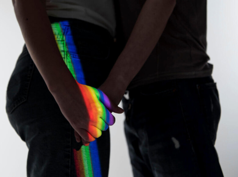 Homophobie Symbolbild: Die Schatten zwei Männer, die sich gegenüberstehend an der Hand halten werden von einem Regenbogen-Licht angestrahlt. Homophobia Symbolic image: The shadows of two men standing opposite each other, holding hands, are illuminated by rainbow light.