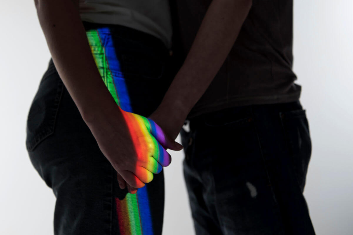 Homophobie Symbolbild: Die Schatten zwei Männer, die sich gegenüberstehend an der Hand halten werden von einem Regenbogen-Licht angestrahlt. Homophobia Symbolic image: The shadows of two men standing opposite each other, holding hands, are illuminated by rainbow light.