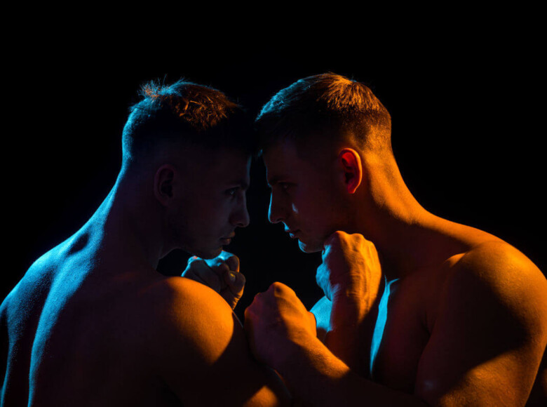 WRAP BDSM Symbolbild: Die nackten Oberkörper zweier Männer, in spärlich orange-farbenes Licht getaucht, die ihre Köpfe aneinanderhalten. . BDSM WRAP Symbolic image: The naked upper bodies of two men, bathed in sparse orange light, holding their heads together.