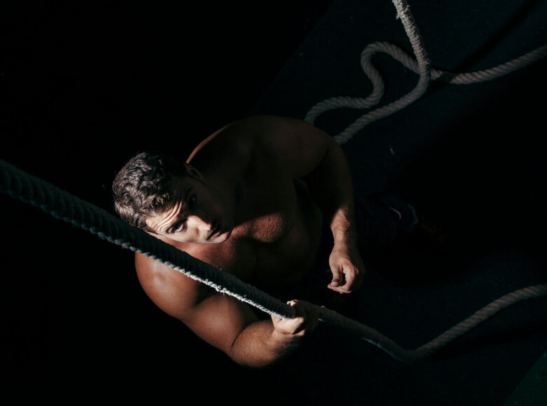 BDSM Shibari Erfahrungsbericht. Symbolbild: Ein Mann sitzt im Dunklen und blickt nach oben in die Kamera. Er ist nackt und mit einem Seil gefesselt. BDSM Shibari experience report. Symbolic image: A man sits in the dark and looks up at the camera. He is naked and tied up with rope.