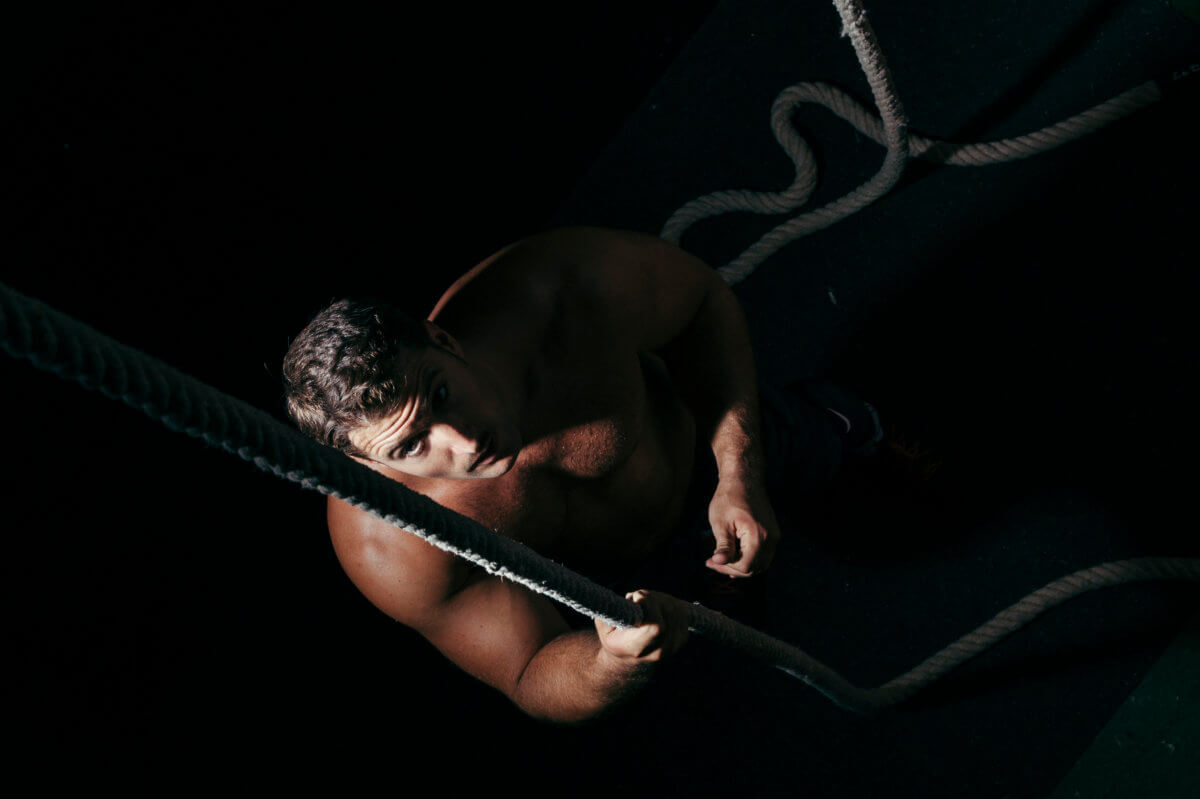 BDSM Shibari Erfahrungsbericht. Symbolbild: Ein Mann sitzt im Dunklen und blickt nach oben in die Kamera. Er ist nackt und mit einem Seil gefesselt. BDSM Shibari experience report. Symbolic image: A man sits in the dark and looks up at the camera. He is naked and tied up with rope.