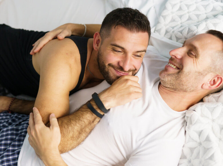 Dry Humping Positionen. Symbolbild: Zwei Männer liegen lachend und sich umarmend auf dem Bett. Englisch: Dry humping positions. Symbolic image: Two men lying on the bed, laughing and hugging each other.
