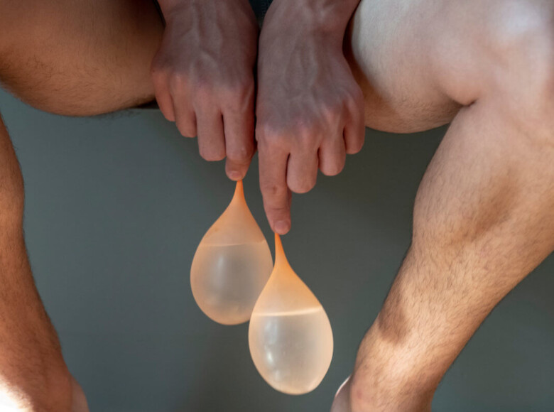 Schlucken oder Spucken Symbolbild: Hände halten zwei mit Wasser gefüllte Ballons. Im Hintergrund nackte Männer-Beine. Swallow or spit Symbolic image: Hands holding two balloons filled with water. Naked men's legs in the background.